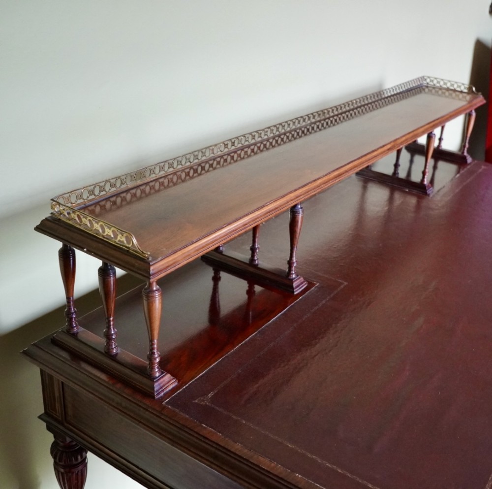 A Victorian rosewood writing desk featuring a brass gallery shelf, three drawers with brass handles, turned fluted legs, and curved stretchers. View XII