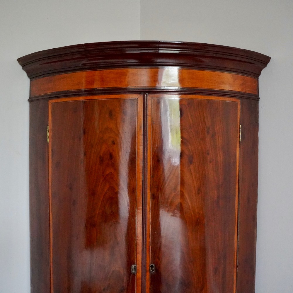 A George III bow front mahogany corner cabinet featuring polished plum pudding veneers, inlaid border stringing, and two curved doors. View II