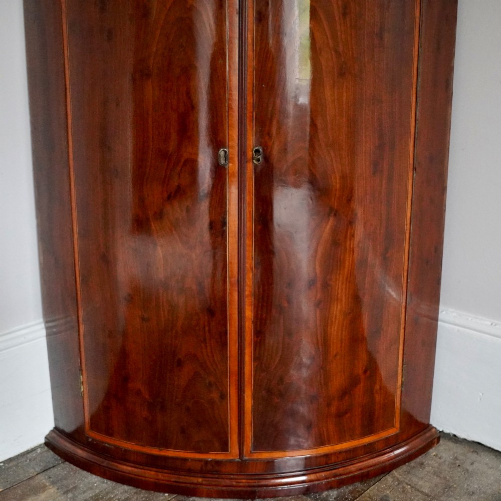 A George III bow front mahogany corner cabinet featuring polished plum pudding veneers, inlaid border stringing, and two curved doors. View III
