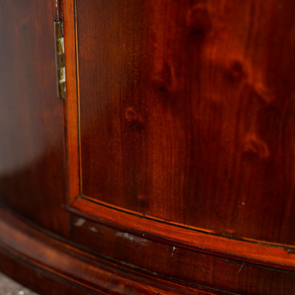 A George III bow front mahogany corner cabinet featuring polished plum pudding veneers, inlaid border stringing, and two curved doors. View IX