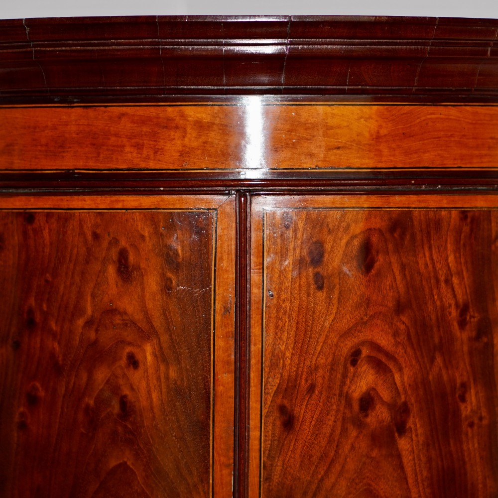 A George III bow front mahogany corner cabinet featuring polished plum pudding veneers, inlaid border stringing, and two curved doors. View X