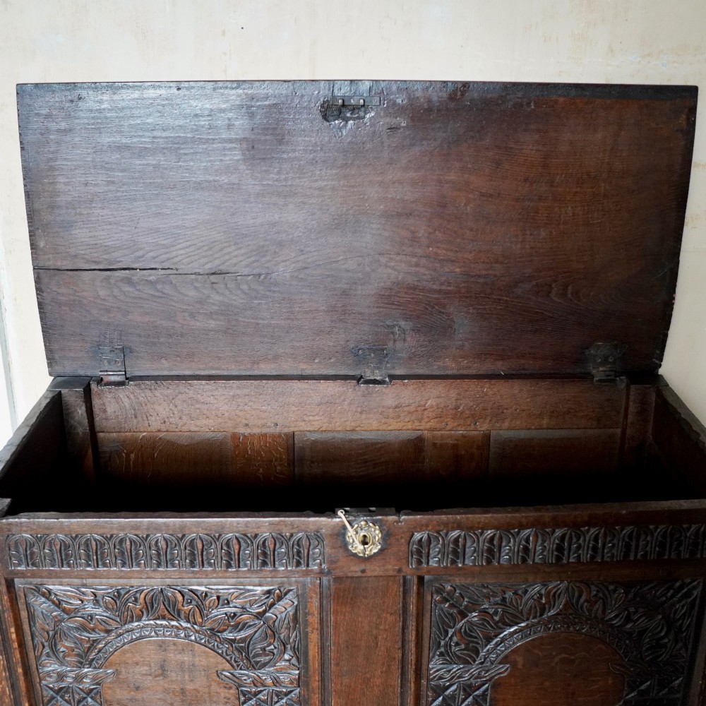 A 17th-century joined oak coffer featuring carved arch front panels, a carved top frieze, hinged lid, and brass keyhole escutcheon. View VI