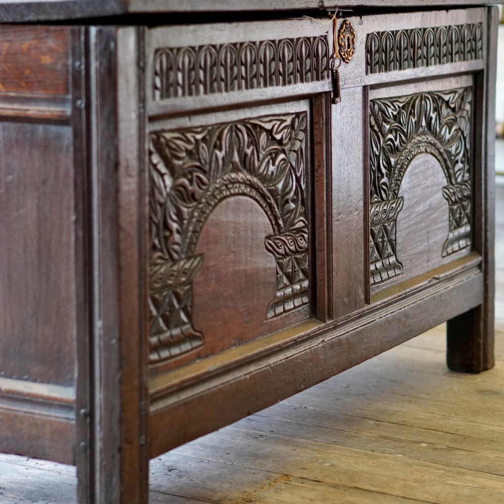 A 17th-century joined oak coffer featuring carved arch front panels, a carved top frieze, hinged lid, and brass keyhole escutcheon. View XIII