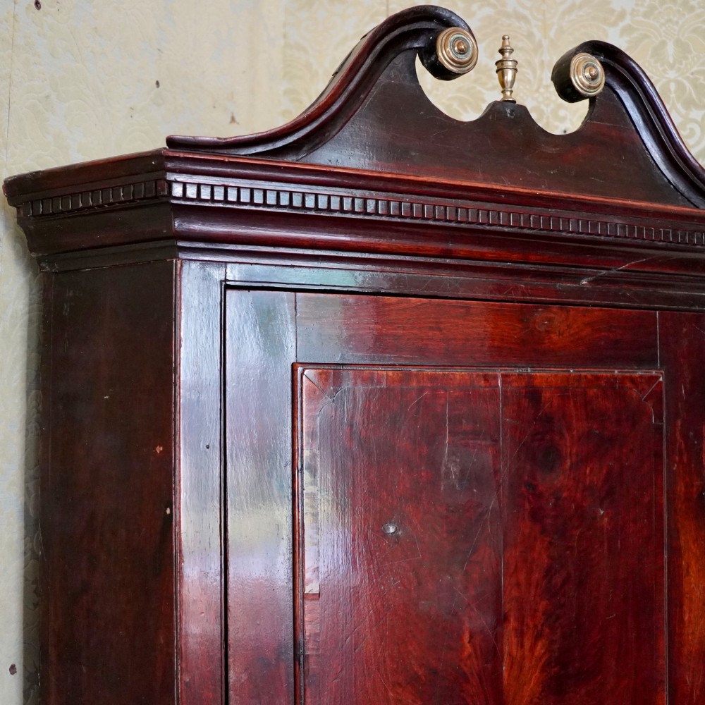 A tall George III standing mahogany corner cupboard featuring a swan-neck pediment with central brass finial and upper and lower fielded panel doors. View VI