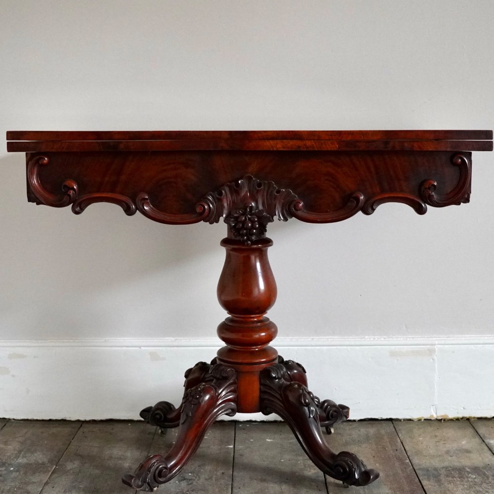 A Victorian figured mahogany card table featuring a turnover top, carved frieze, turned pedestal, and scroll legs with castors. View II
