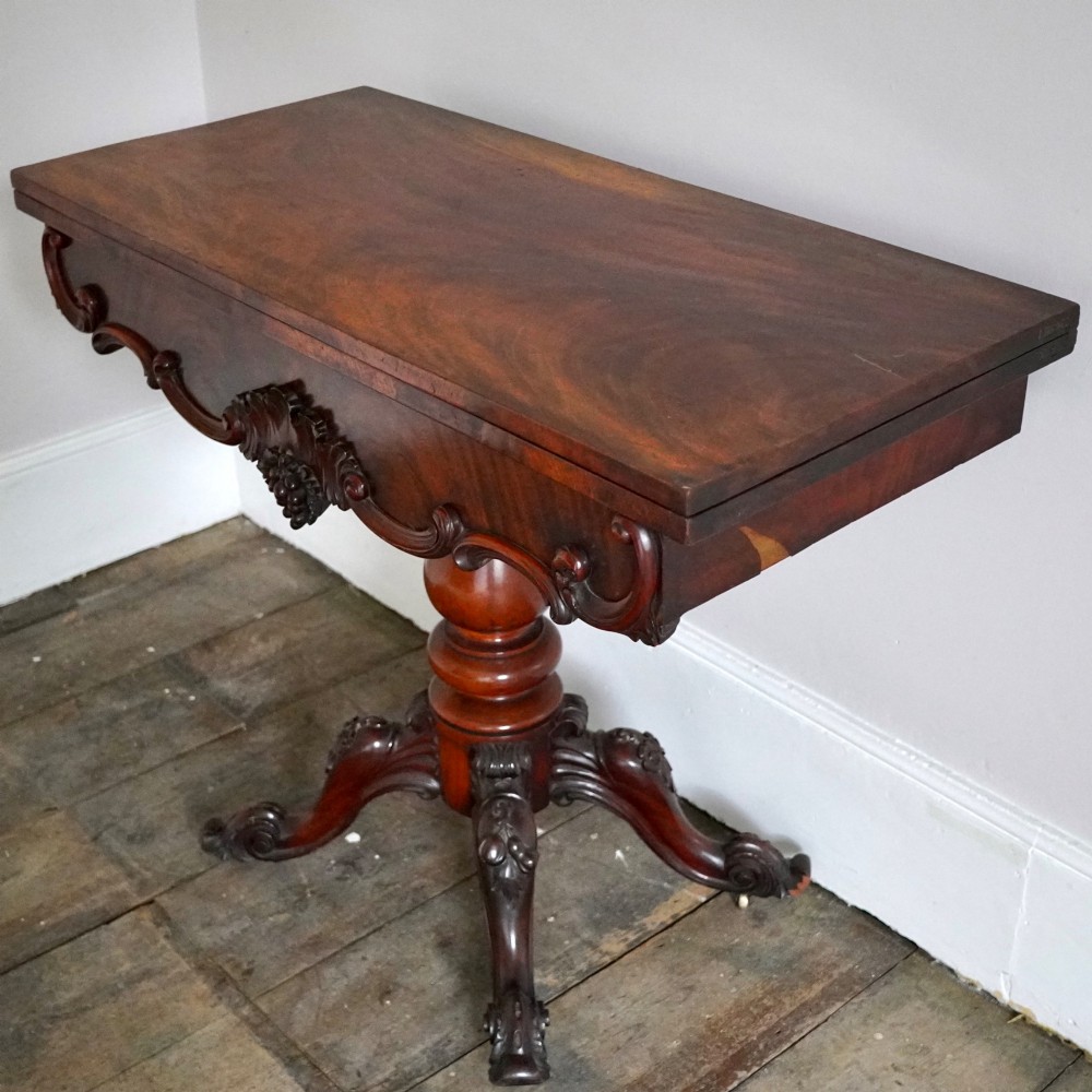 A Victorian figured mahogany card table featuring a turnover top, carved frieze, turned pedestal, and scroll legs with castors. View IV