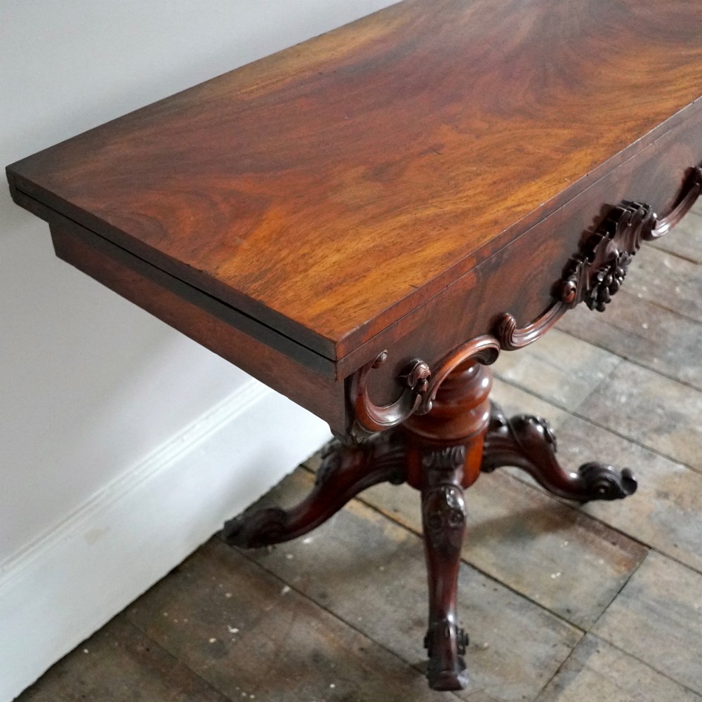 A Victorian figured mahogany card table featuring a turnover top, carved frieze, turned pedestal, and scroll legs with castors. View V