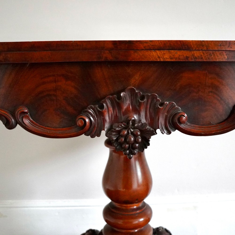 A Victorian figured mahogany card table featuring a turnover top, carved frieze, turned pedestal, and scroll legs with castors. View VI