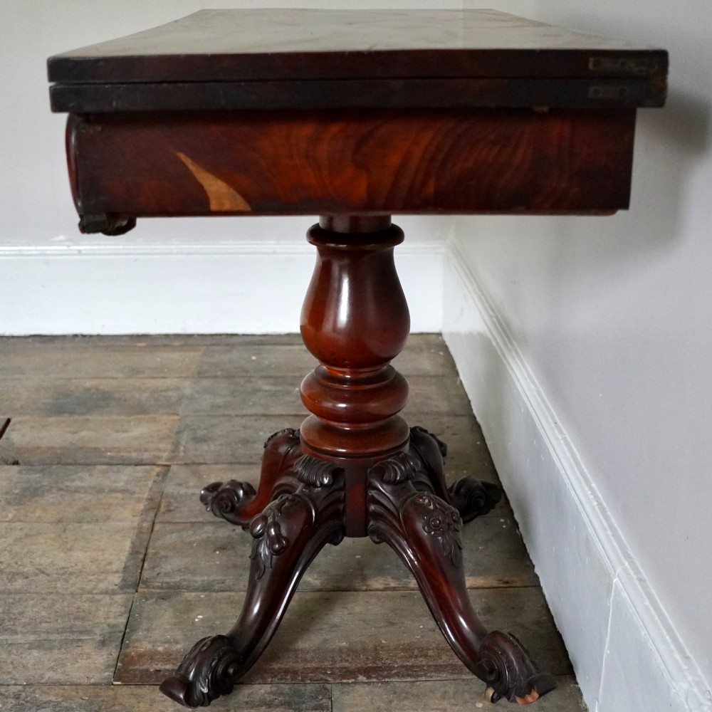 A Victorian figured mahogany card table featuring a turnover top, carved frieze, turned pedestal, and scroll legs with castors. View VIII