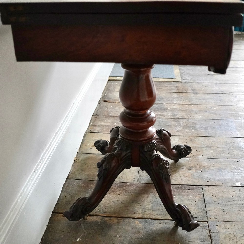 A Victorian figured mahogany card table featuring a turnover top, carved frieze, turned pedestal, and scroll legs with castors. View IX