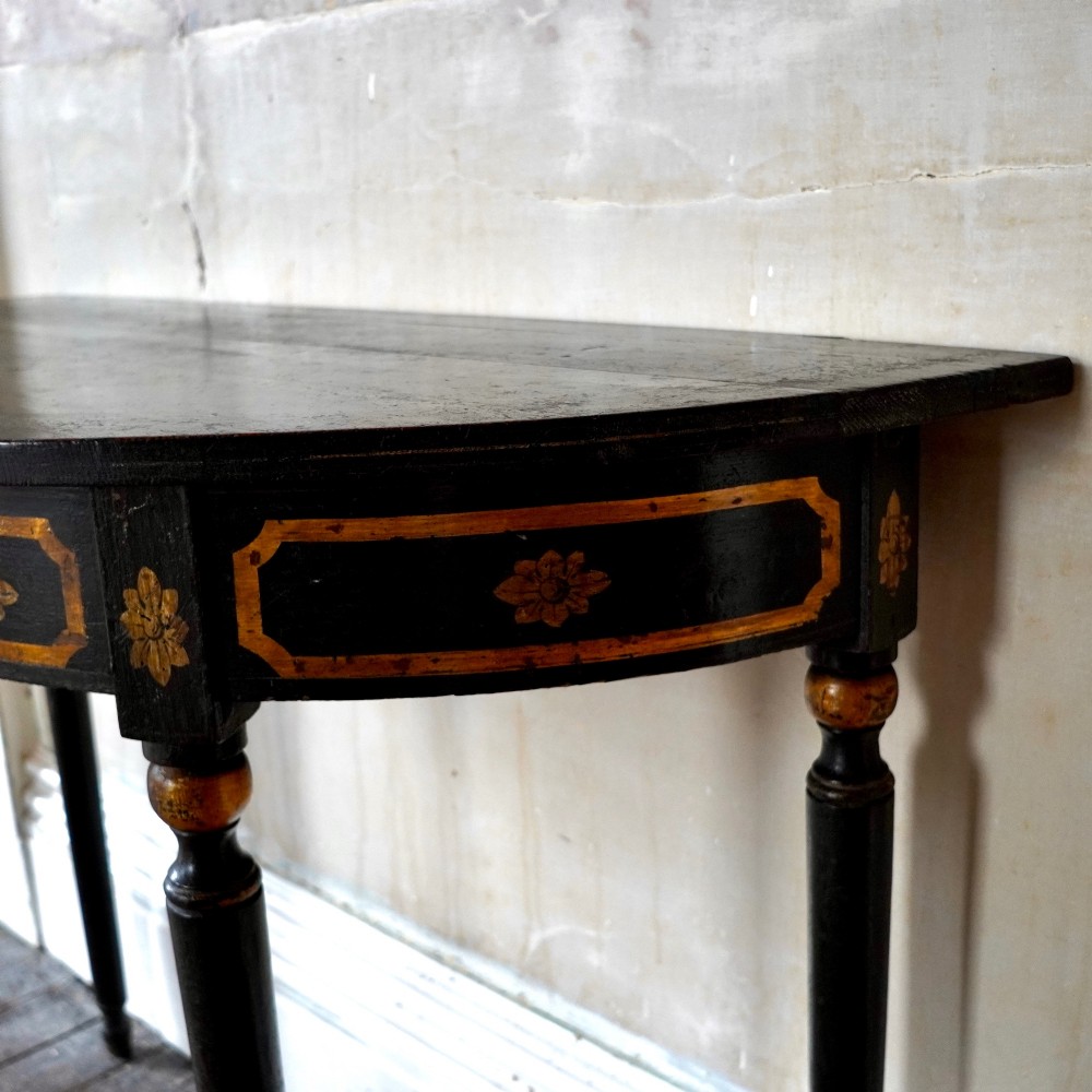 A Regency period semi-circular console table painted black with gold-gilded classical panel motifs and four slender turned legs. View X