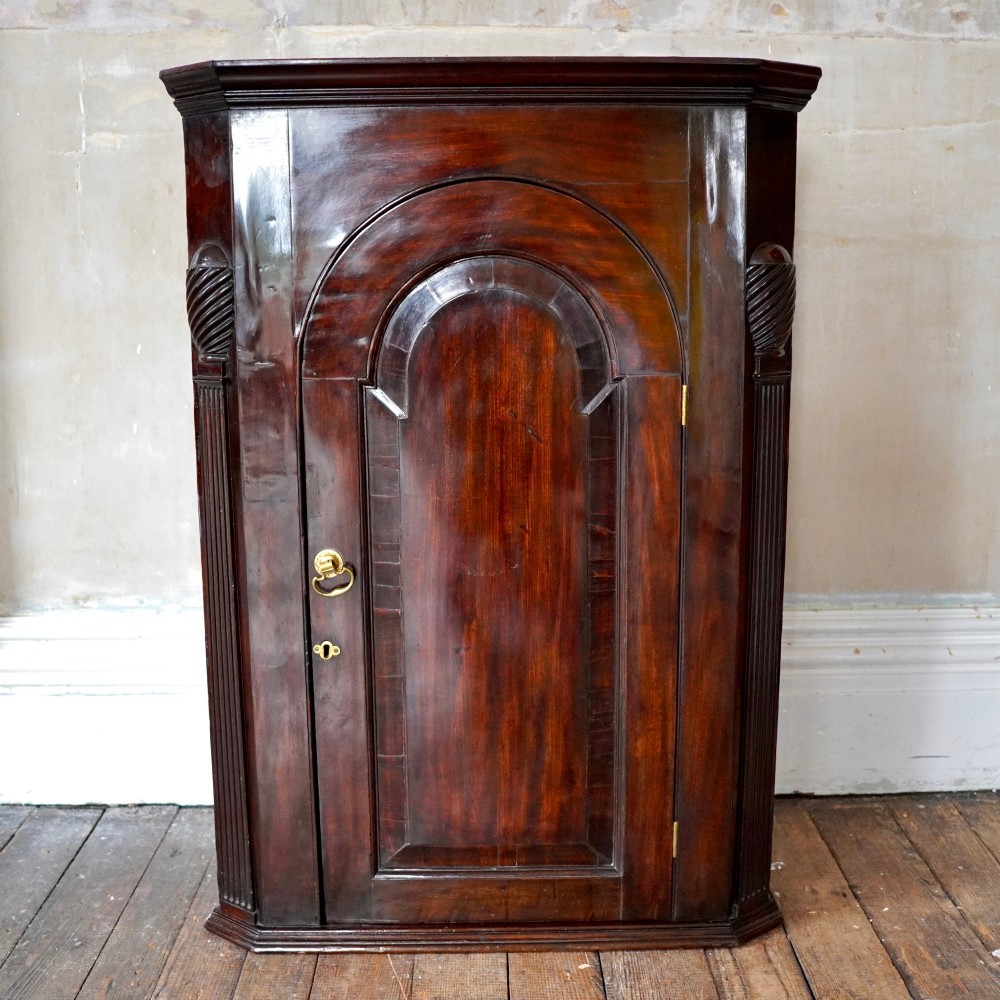 Mid-18th century Georgian mahogany hanging corner cupboard featuring an arched panelled door, brass hardware, and fluted pilasters. View III