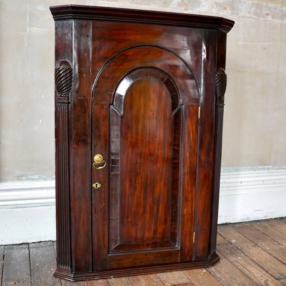 Mid-18th century Georgian mahogany hanging corner cupboard featuring an arched panelled door, brass hardware, and fluted pilasters. View II