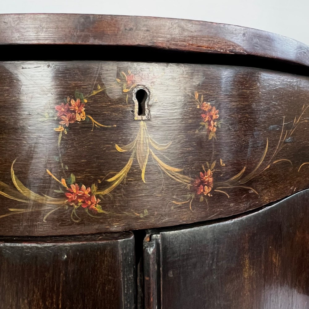 A dark mahogany serpentine-front side cabinet with hand-painted floral swags and figurative door panel vignettes resting on slender square tapered legs. View V