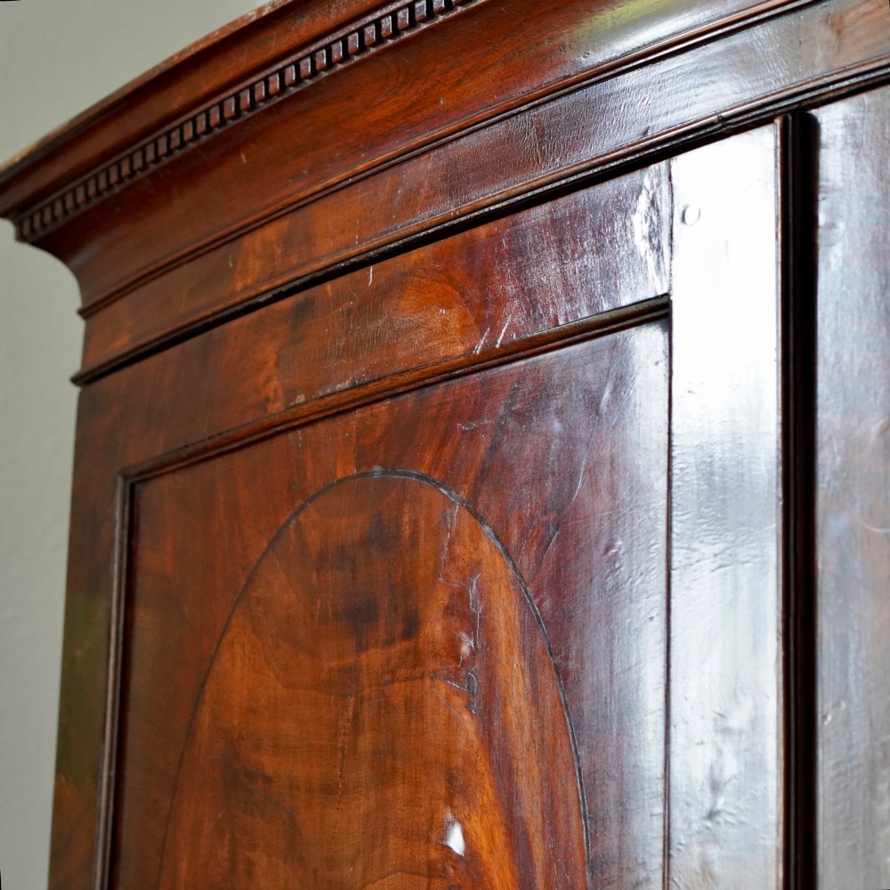 A George III bowfront mahogany wardrobe featuring twin panelled doors with oval flame mahogany veneers above two drawers and flared bracket feet. View III