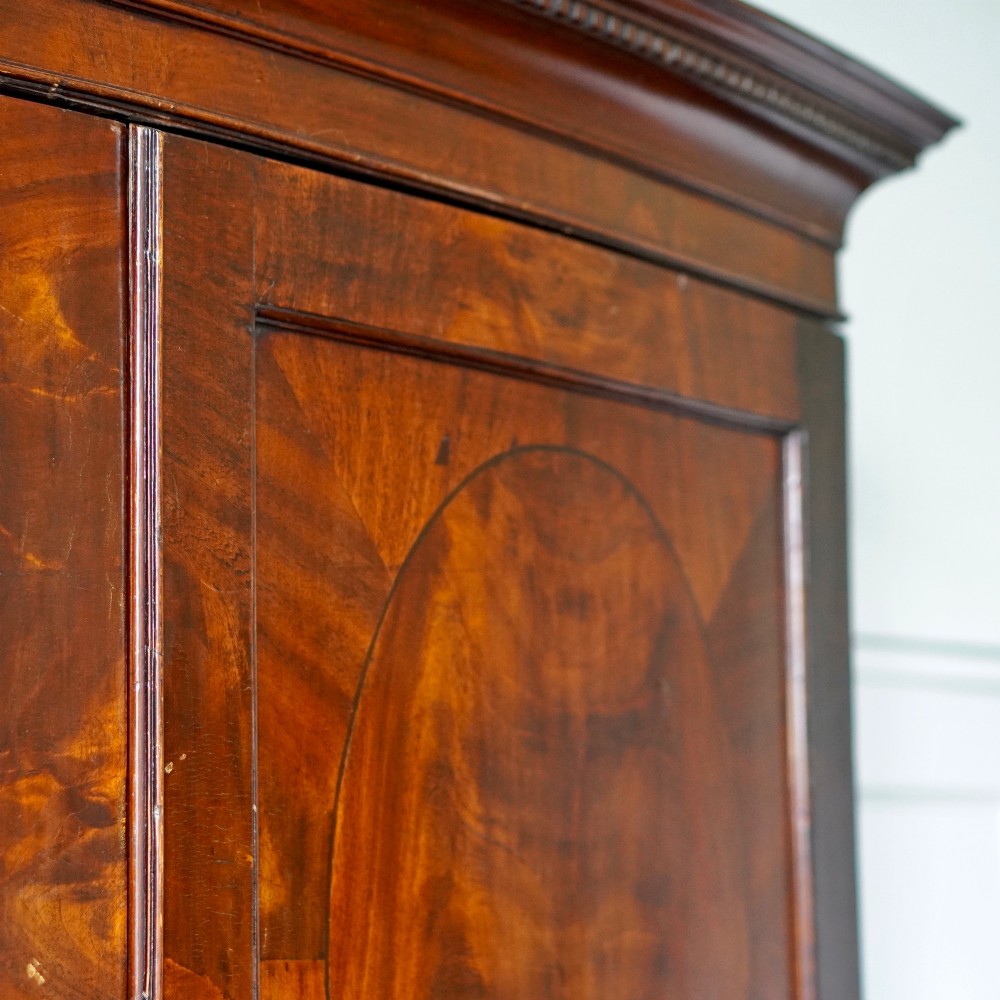 A George III bowfront mahogany wardrobe featuring twin panelled doors with oval flame mahogany veneers above two drawers and flared bracket feet. View IV