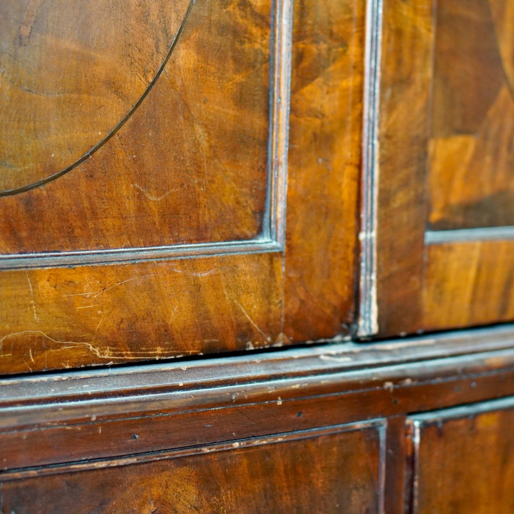 A George III bowfront mahogany wardrobe featuring twin panelled doors with oval flame mahogany veneers above two drawers and flared bracket feet. View VII