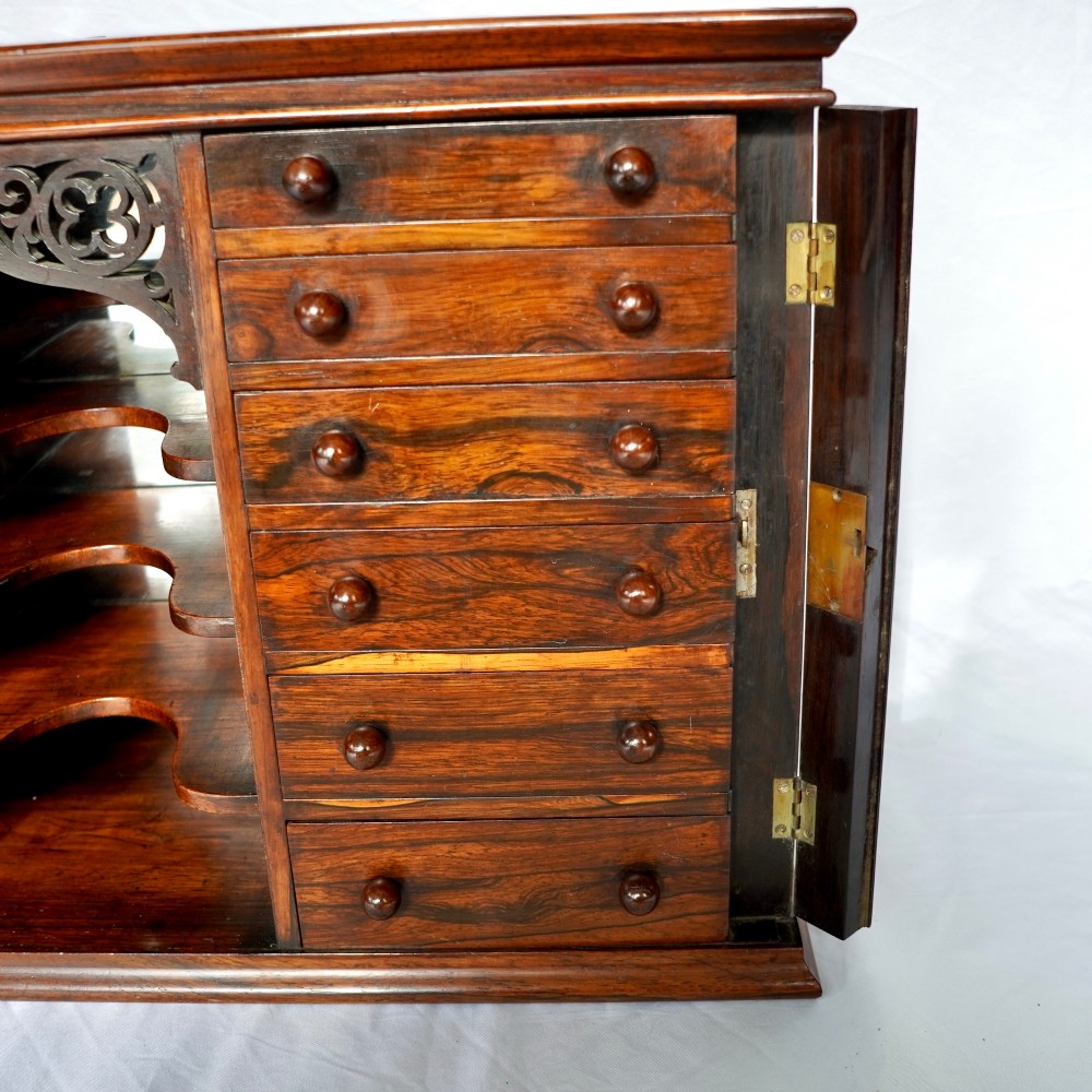 A Regency period dark rosewood tabletop collectors cabinet featuring twin banks of small drawers flanking a central mirrored shelf unit with Gothic arched carving. View X