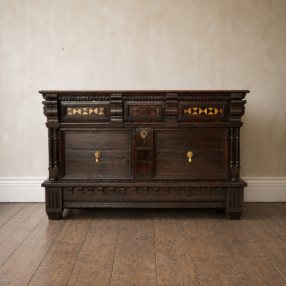A dark oak 17th-century style chest of drawers featuring geometric timber inlay along the top frieze, applied mouldings, and brass drop handles.