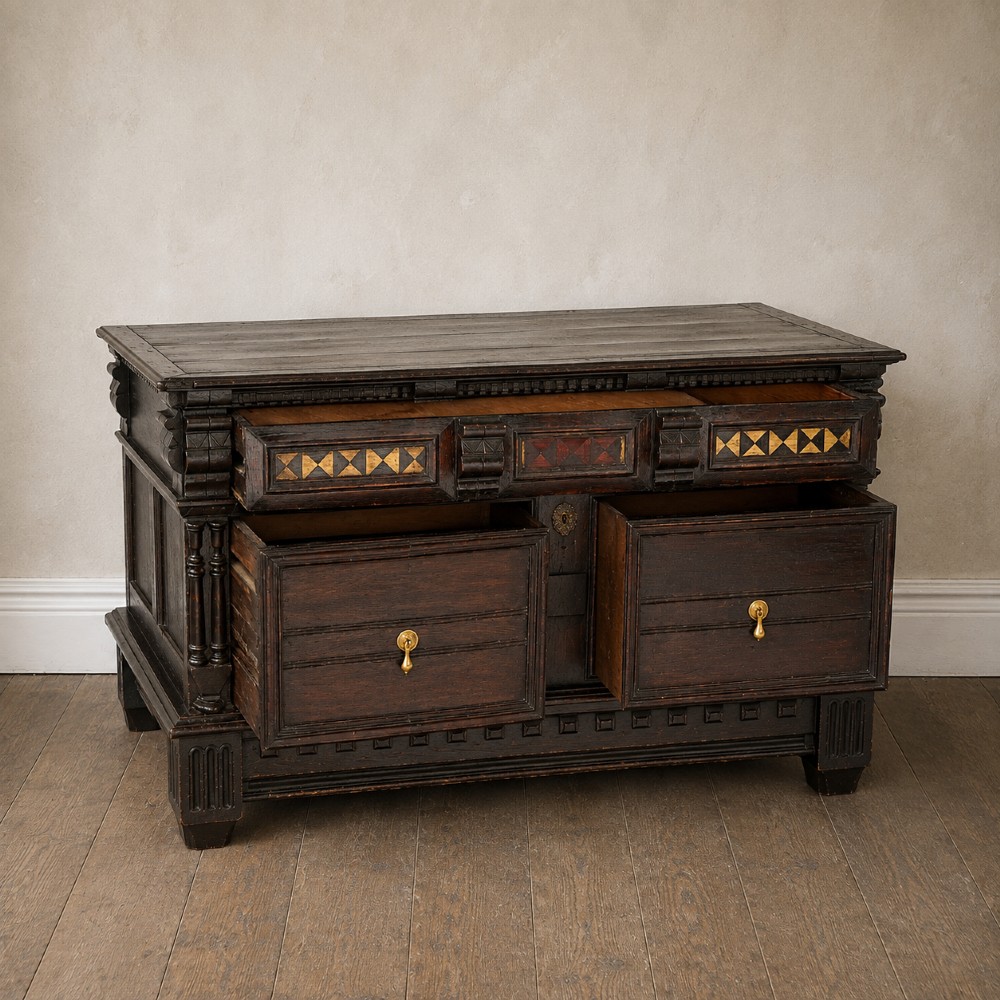 A dark oak 17th-century style chest of drawers featuring geometric timber inlay along the top frieze, applied mouldings, and brass drop handles. View II