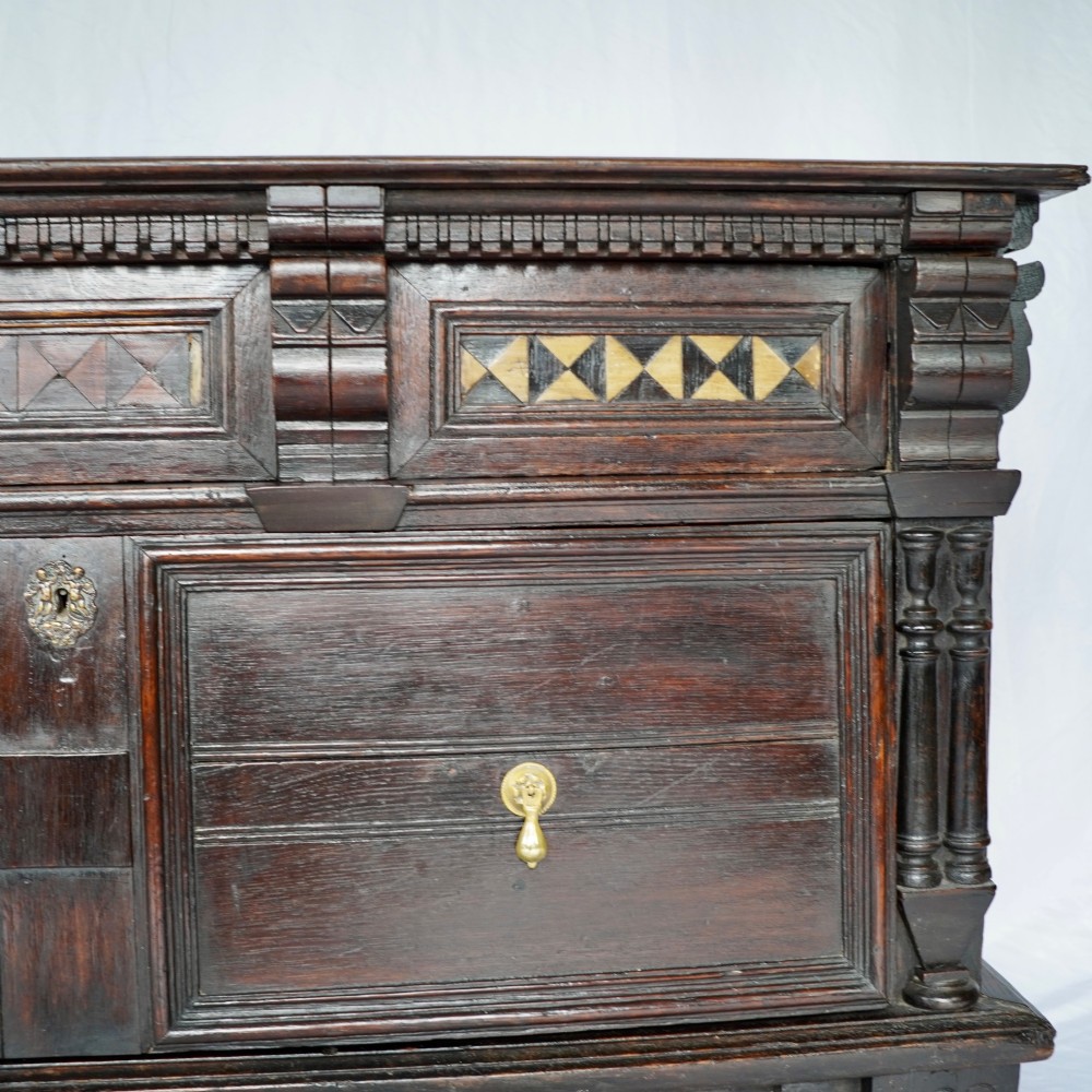 A dark oak 17th-century style chest of drawers featuring geometric timber inlay along the top frieze, applied mouldings, and brass drop handles. View IX