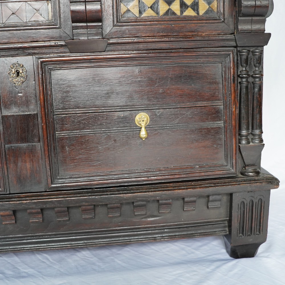 A dark oak 17th-century style chest of drawers featuring geometric timber inlay along the top frieze, applied mouldings, and brass drop handles. View X
