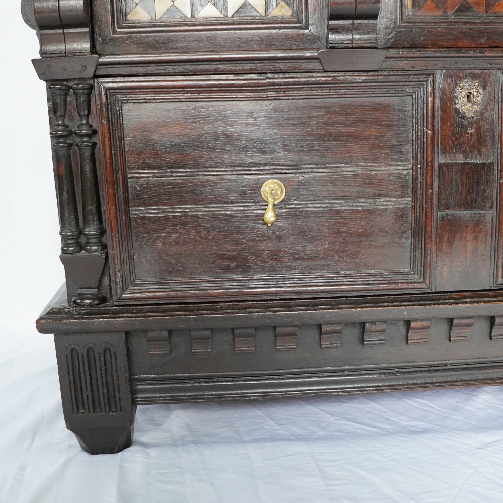 A dark oak 17th-century style chest of drawers featuring geometric timber inlay along the top frieze, applied mouldings, and brass drop handles. View XIII