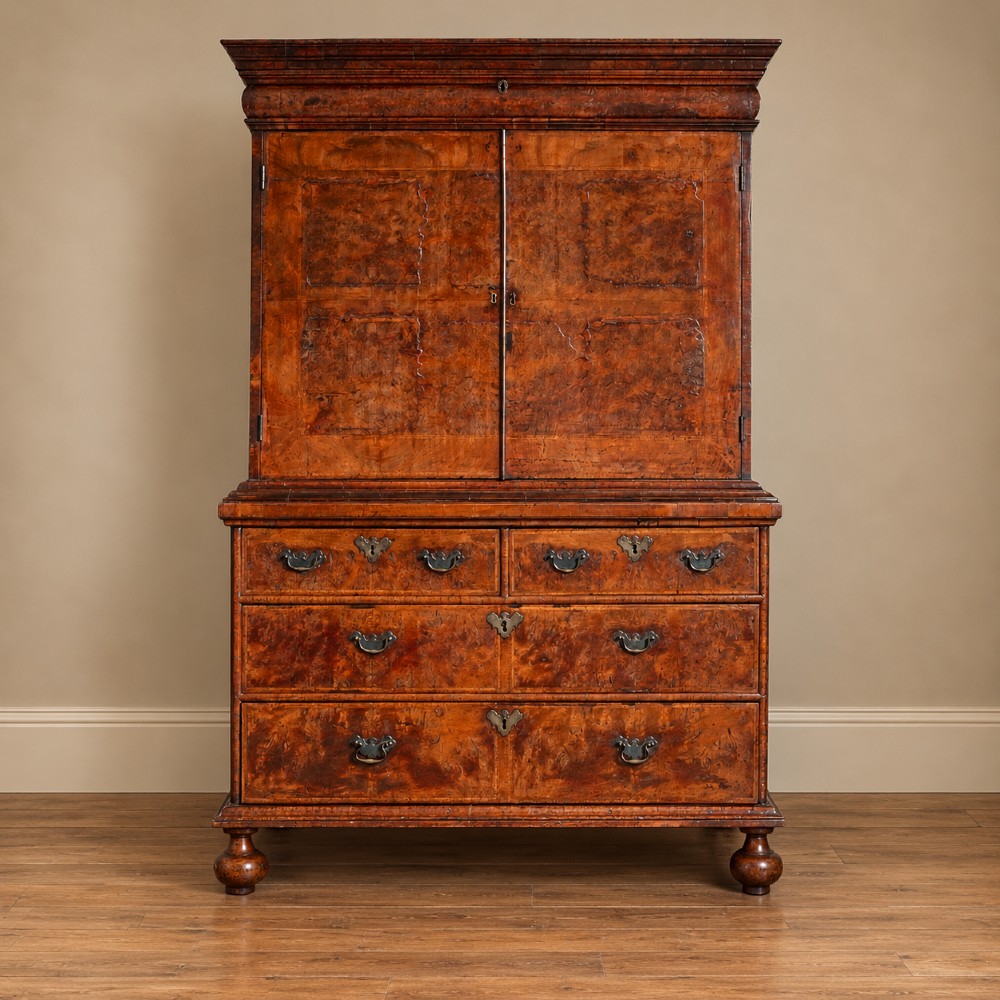 An antique Queen Anne burr walnut linen press featuring upper double cupboard doors over three lower drawers with brass handles on turned bun feet.