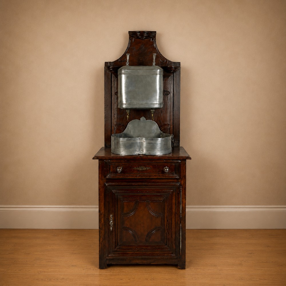 An 18th-century dark chestnut wood lavabo cabinet featuring a carved backboard mounted with a pewter cistern above a matching pewter basin.