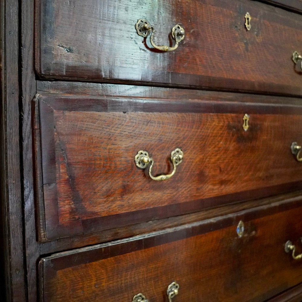 A Georgian dark oak chest of drawers featuring two short drawers over three long drawers with mahogany crossbanding and brass swan-neck handles. View VII