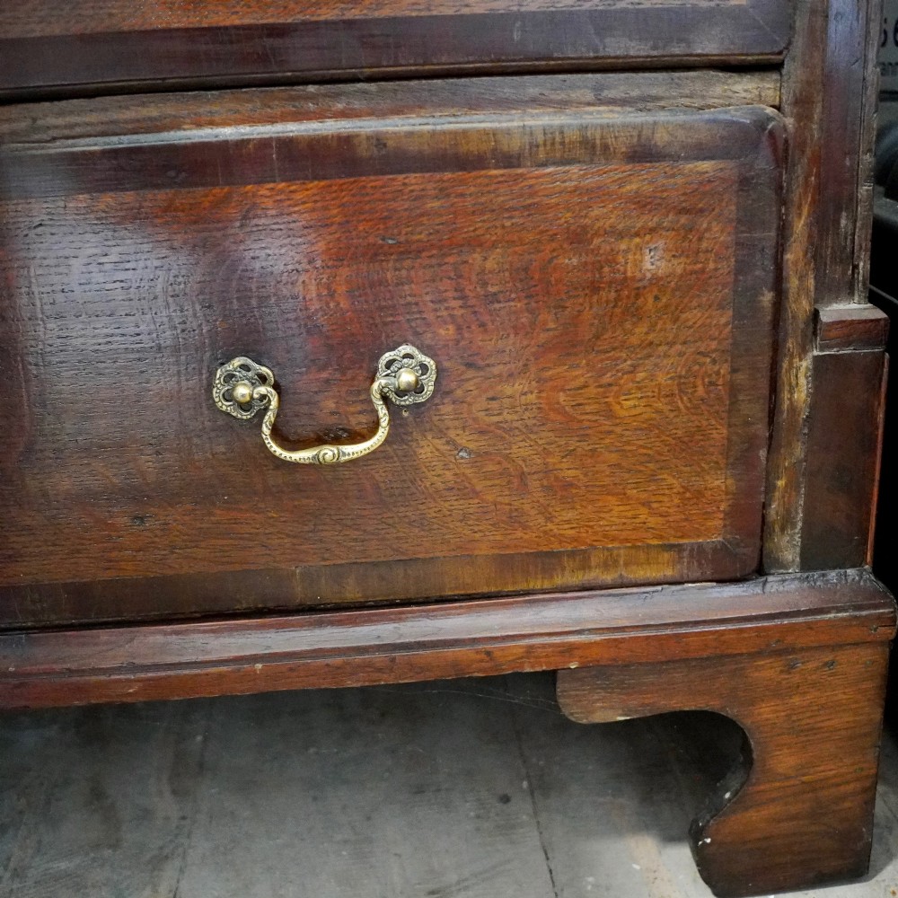 A Georgian dark oak chest of drawers featuring two short drawers over three long drawers with mahogany crossbanding and brass swan-neck handles. View IX
