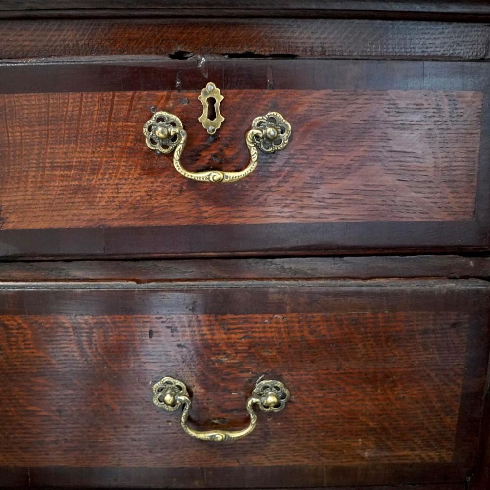 A Georgian dark oak chest of drawers featuring two short drawers over three long drawers with mahogany crossbanding and brass swan-neck handles. View X