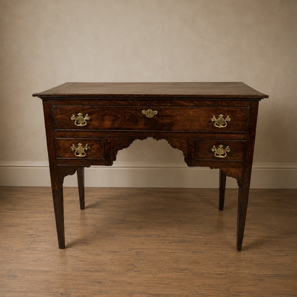 An 18th-century Georgian dark oak lowboy side table featuring three drawers with brass handles, a shaped apron, and slender square legs.