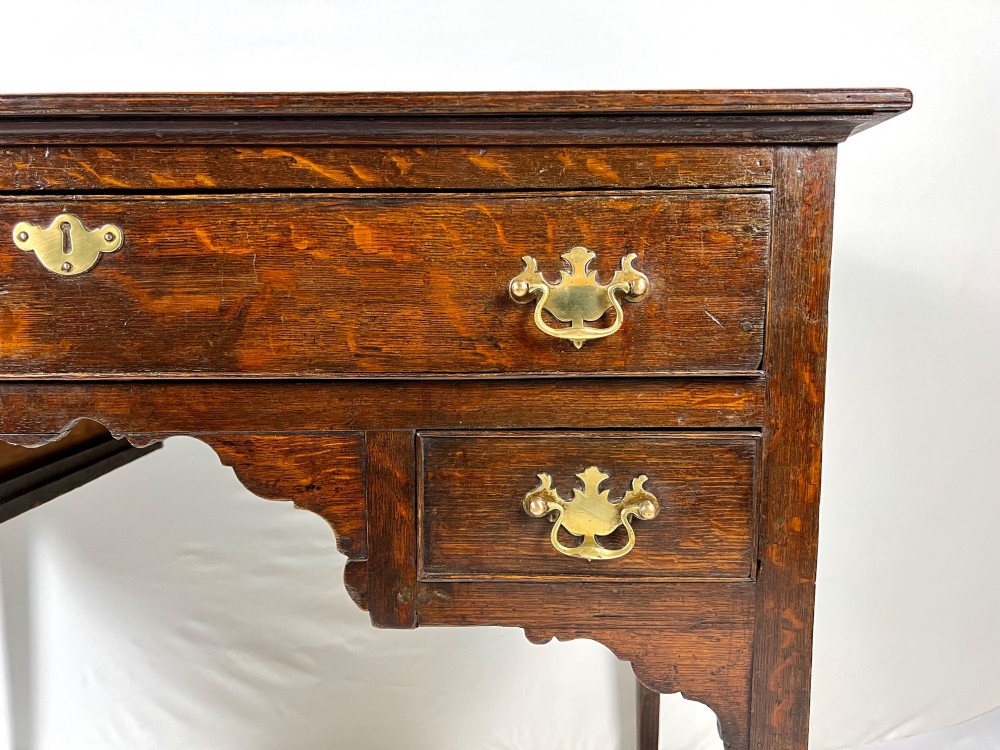 An 18th-century Georgian dark oak lowboy side table featuring three drawers with brass handles, a shaped apron, and slender square legs. View X
