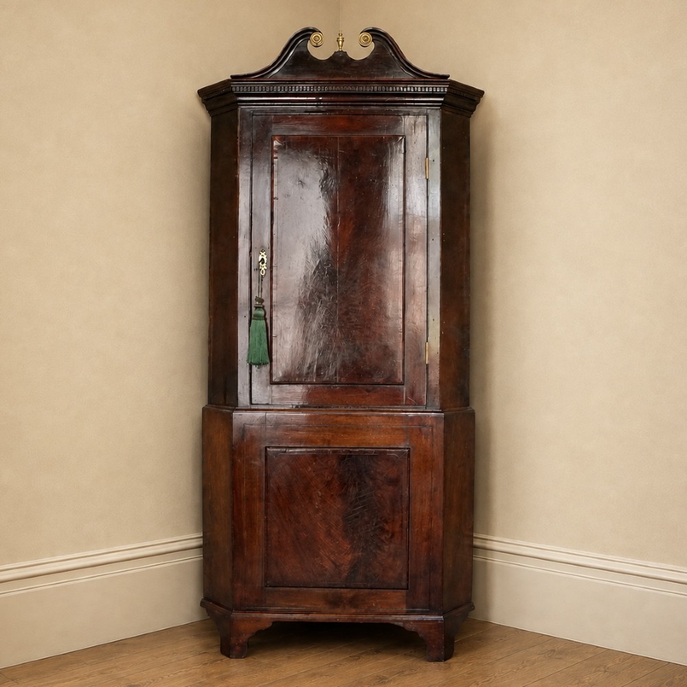 A tall George III standing mahogany corner cupboard featuring a swan-neck pediment with central brass finial and upper and lower fielded panel doors.