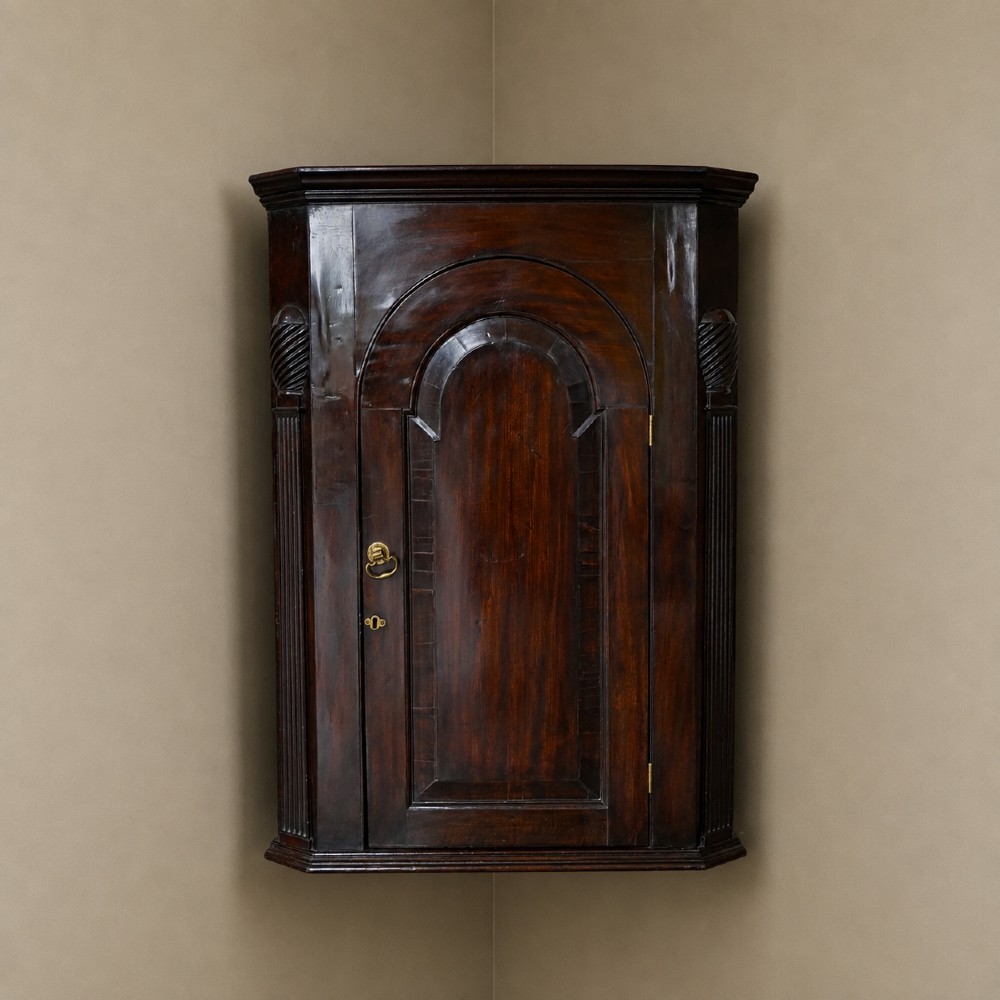 Mid-18th century Georgian mahogany hanging corner cupboard featuring an arched panelled door, brass hardware, and fluted pilasters.