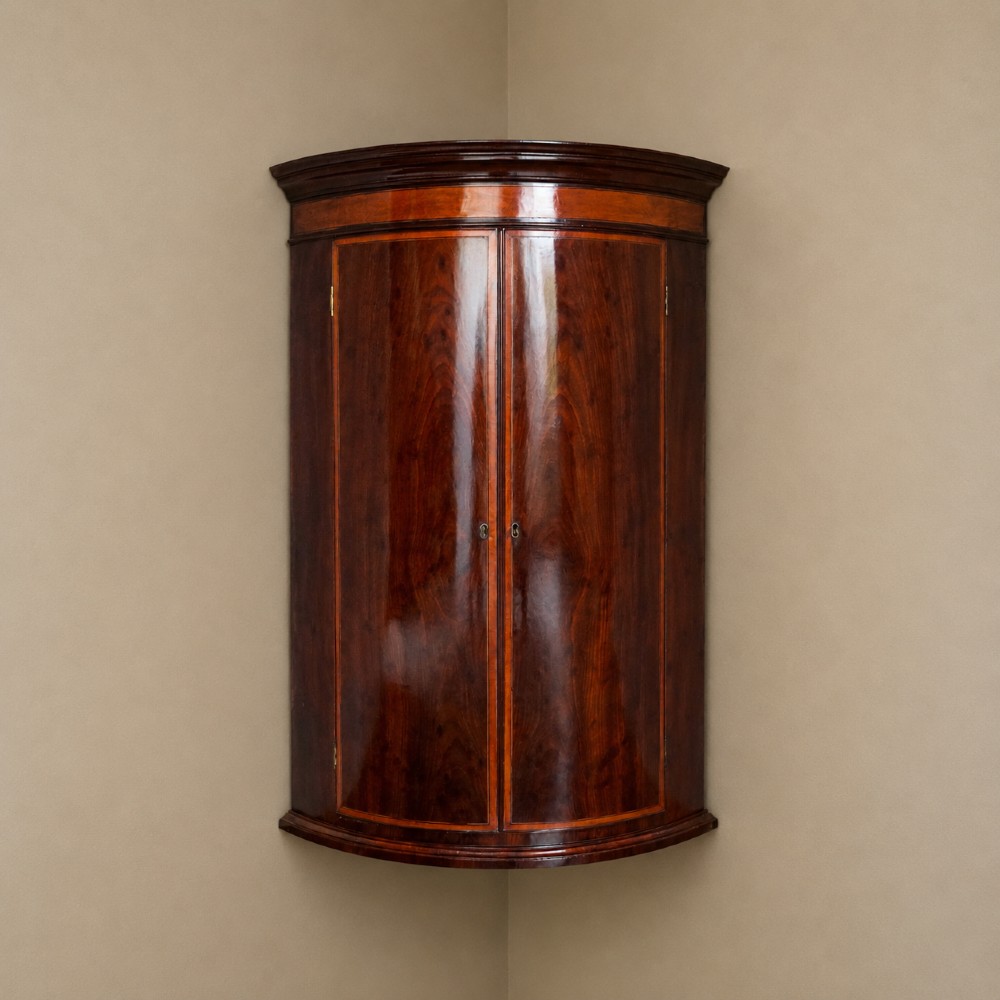A George III bow front mahogany corner cabinet featuring polished plum pudding veneers, inlaid border stringing, and two curved doors.