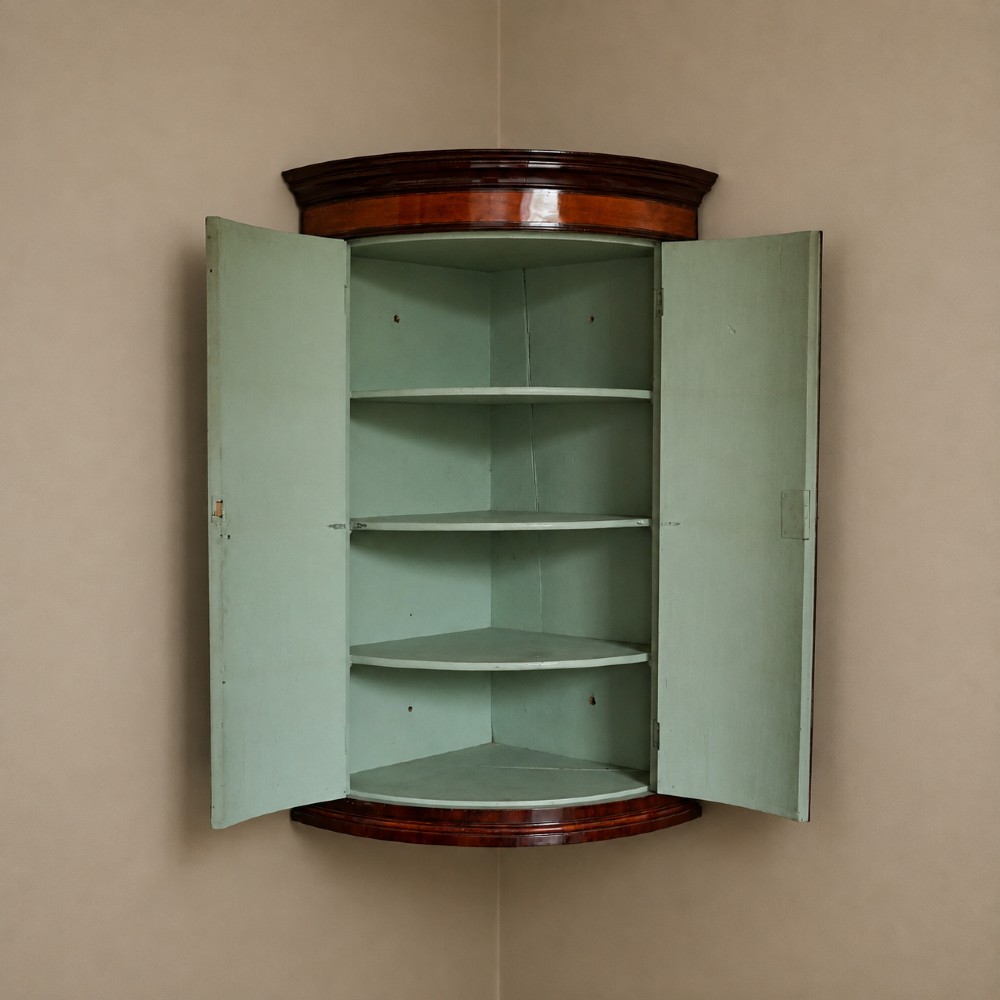 A George III bow front mahogany corner cabinet featuring polished plum pudding veneers, inlaid border stringing, and two curved doors. View XIV