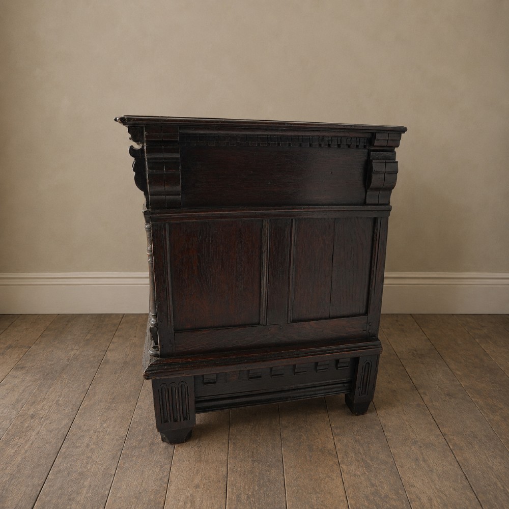 A dark oak 17th-century style chest of drawers featuring geometric timber inlay along the top frieze, applied mouldings, and brass drop handles. View XV