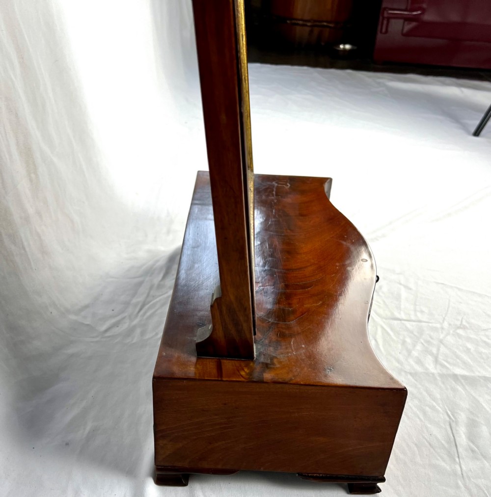 A George III mahogany toilet dressing mirror featuring an upright rectangular swing mirror flanked by brass finials, mounted on a serpentine-front base with three small drawers. View II