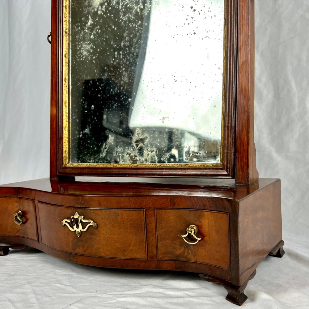 A George III mahogany toilet dressing mirror featuring an upright rectangular swing mirror flanked by brass finials, mounted on a serpentine-front base with three small drawers. View V