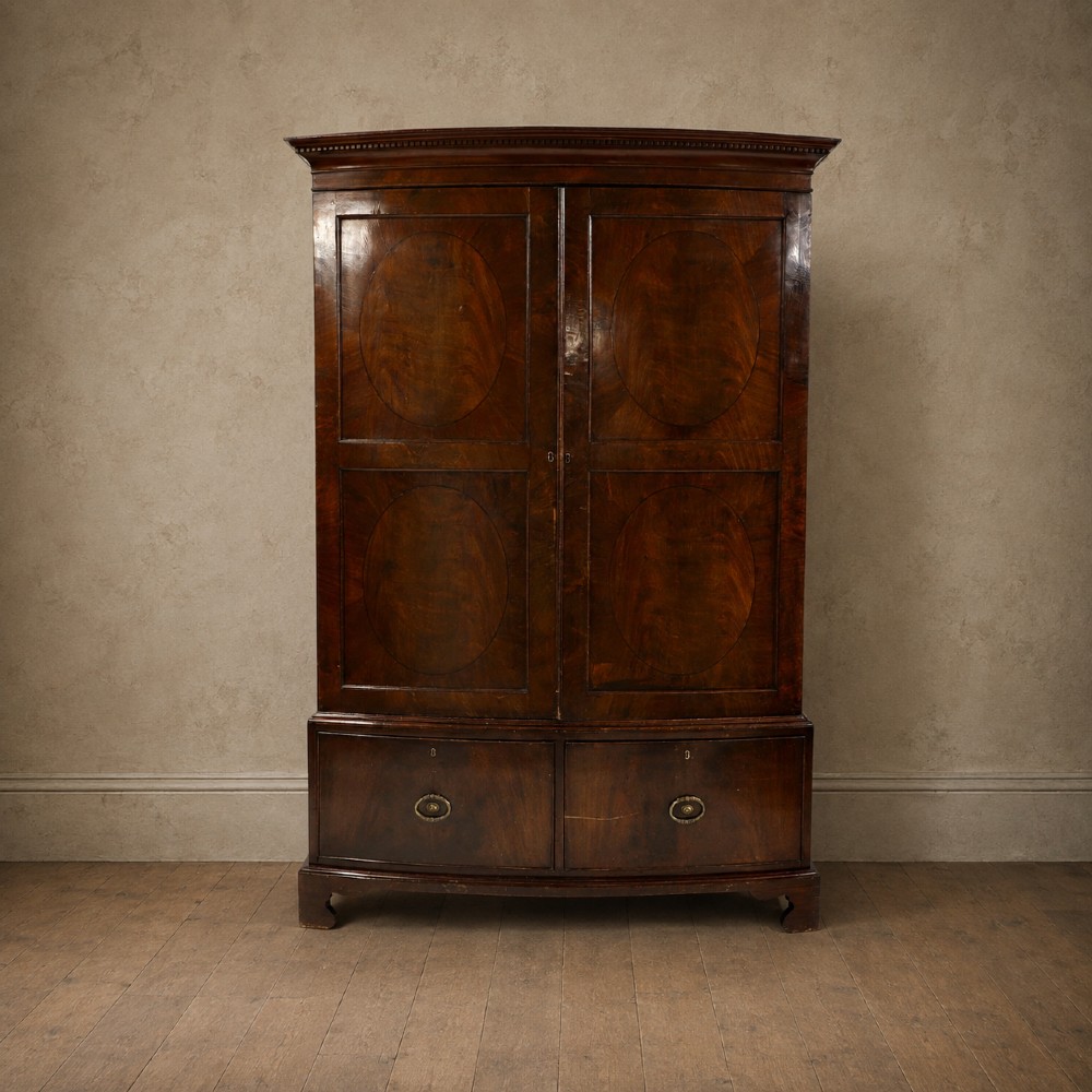 A George III bowfront mahogany wardrobe featuring twin panelled doors with oval flame mahogany veneers above two drawers and flared bracket feet.