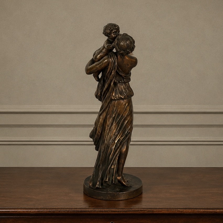 A patinated dark bronze sculpture depicting a draped classical woman holding a small child against her face, set upon a rounded bronze base. View IX