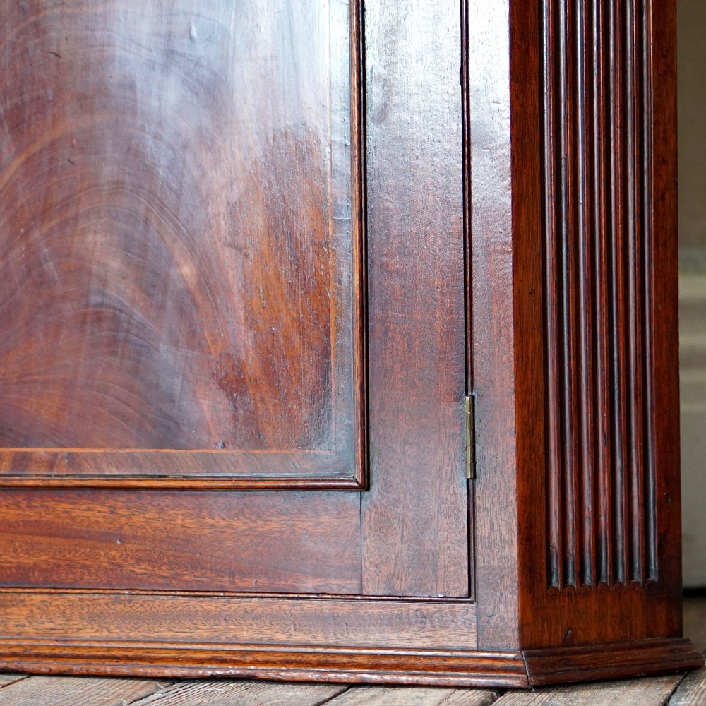 A Georgian dark mahogany hanging corner cupboard featuring a swan-neck pediment with brass paterae and a single panelled door with a brass escutcheon. View XII