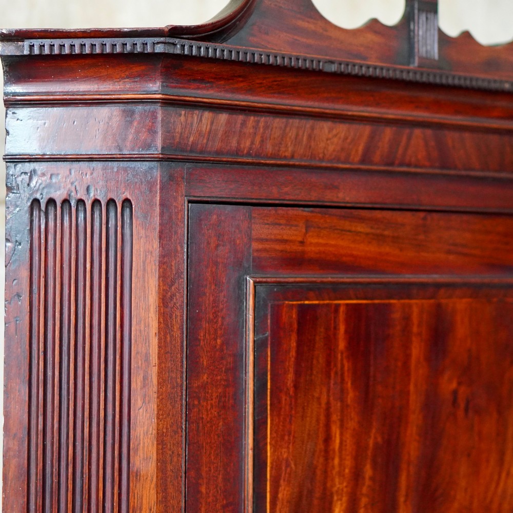 A Georgian dark mahogany hanging corner cupboard featuring a swan-neck pediment with brass paterae and a single panelled door with a brass escutcheon. View XIV