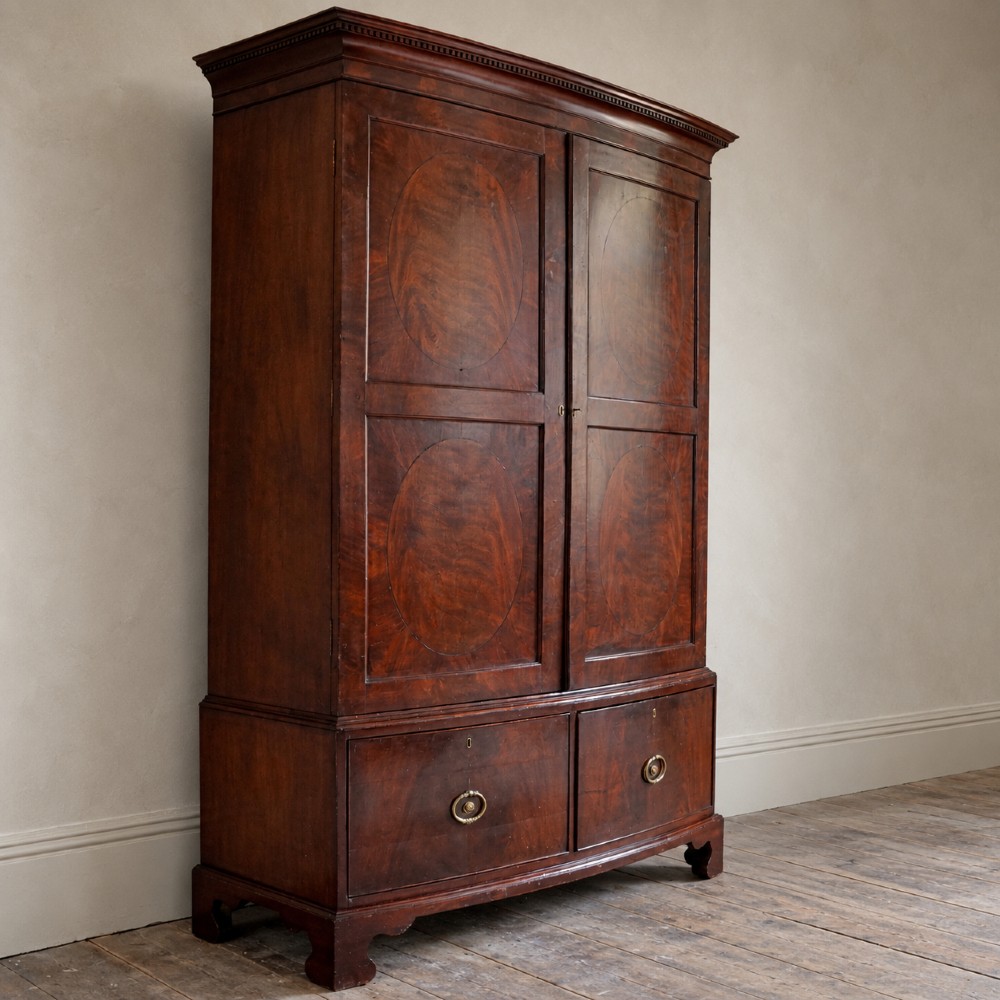 A George III bowfront mahogany wardrobe featuring twin panelled doors with oval flame mahogany veneers above two drawers and flared bracket feet. View III