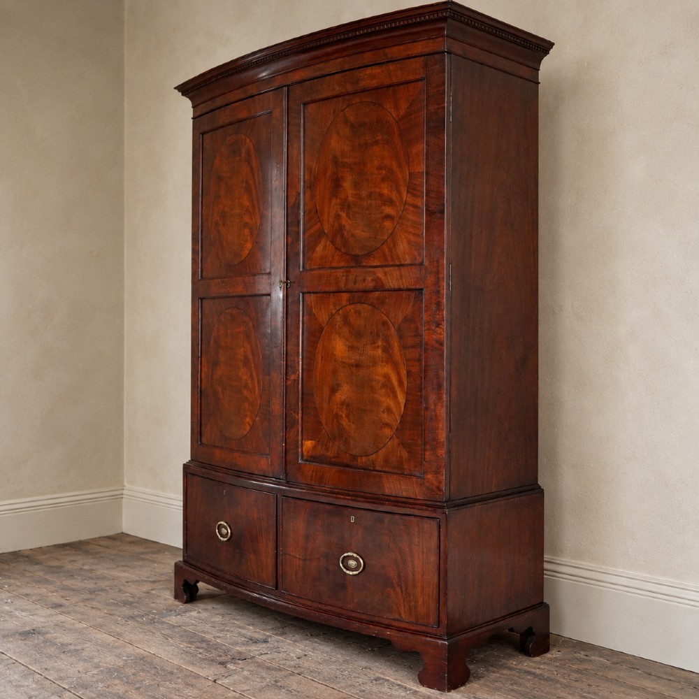 A George III bowfront mahogany wardrobe featuring twin panelled doors with oval flame mahogany veneers above two drawers and flared bracket feet. View IV