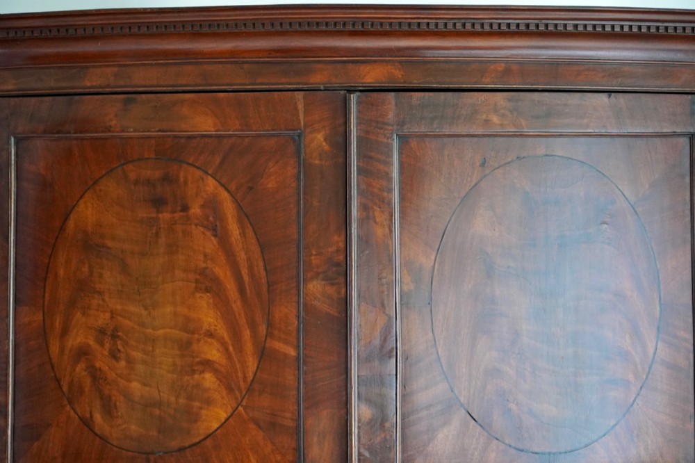 A George III bowfront mahogany wardrobe featuring twin panelled doors with oval flame mahogany veneers above two drawers and flared bracket feet. View XII