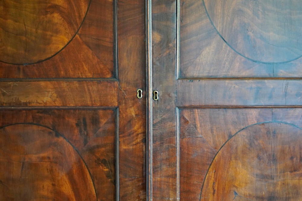A George III bowfront mahogany wardrobe featuring twin panelled doors with oval flame mahogany veneers above two drawers and flared bracket feet. View XIII