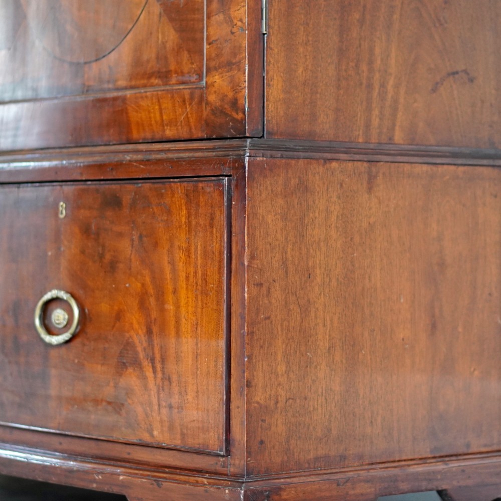 A George III bowfront mahogany wardrobe featuring twin panelled doors with oval flame mahogany veneers above two drawers and flared bracket feet. View XIV